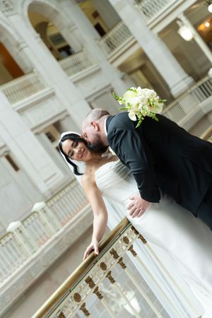 Bride and groom share romantic moment leaning on brass railing in elegant neoclassical wedding venue.