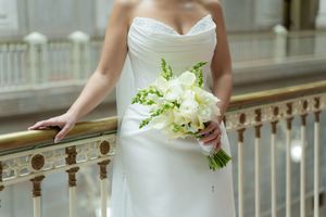Bride in white strapless wedding gown holding elegant white floral bouquet on ornate golden railing staircase.