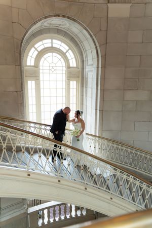 Bride and groom share a romantic moment on a grand curved staircase in a stunning white marble wedding venue.
