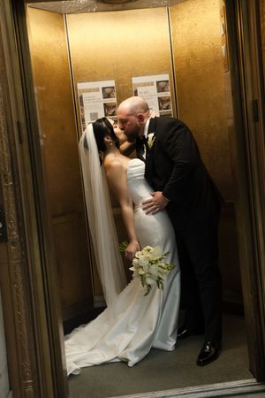 Bride and groom sharing a romantic kiss inside a golden elevator, bride holding white bouquet in wedding dress with veil.