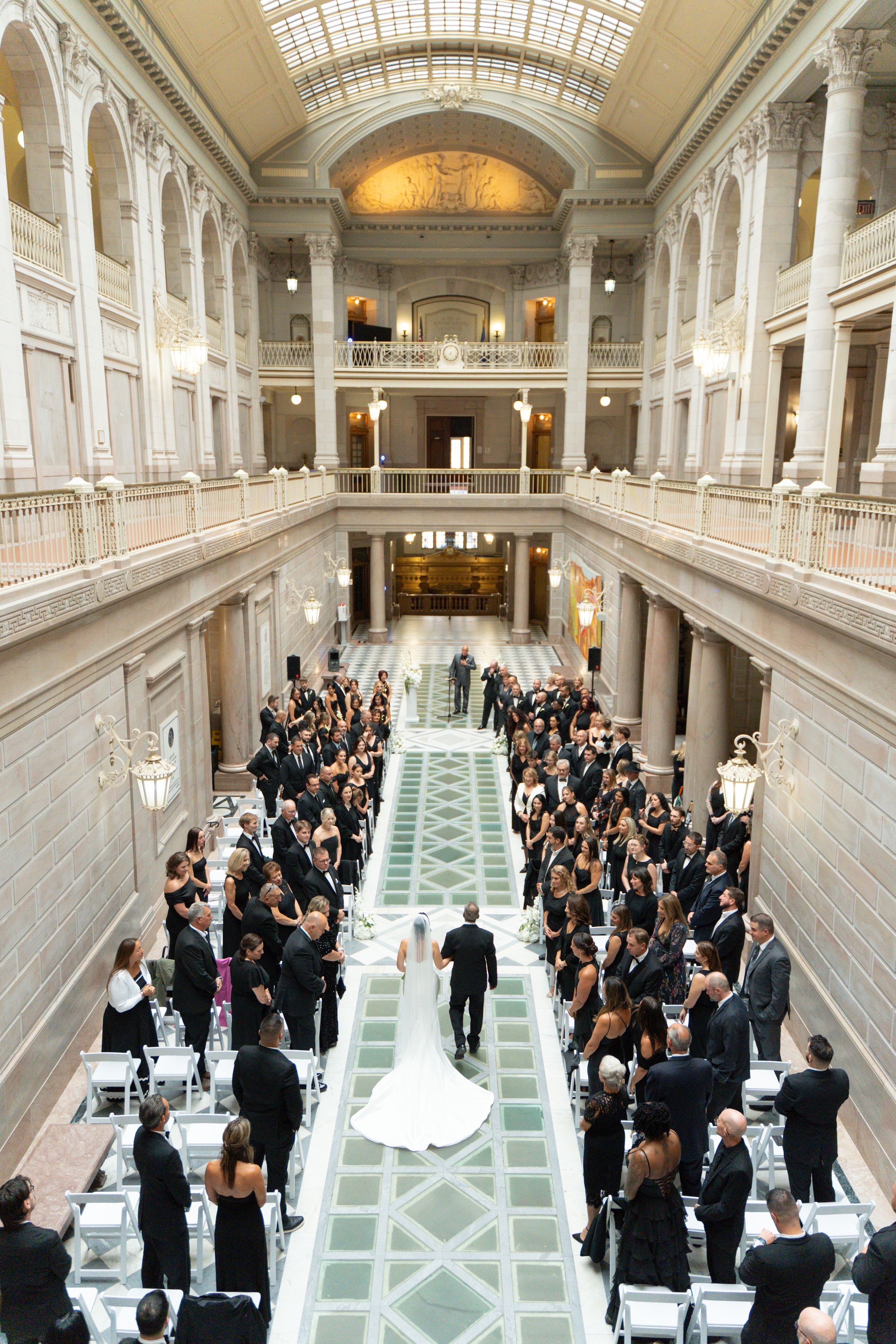 Aerial view of wedding ceremony in grand neoclassical hall with guests in black attire lining aisle.