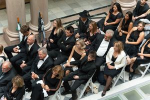 Large group of people dressed in black formal attire seated at an elegant indoor ceremony or funeral service.