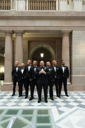 Groom and groomsmen in black tuxedos pose in an elegant marble hall with ornate columns.