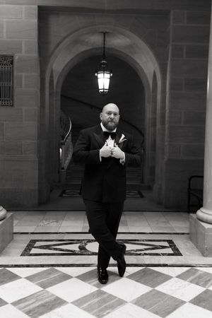 Dapper groom in black tuxedo and bow tie poses in black and white photo under stone archway.