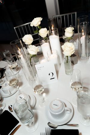 Elegant wedding reception table setting with white roses, pillar candles, crystal glassware, and a table number card.
