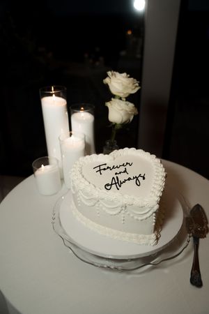 Heart-shaped white wedding cake inscribed Forever and Always surrounded by lit candles on table.