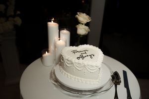 White heart-shaped wedding cake with cursive writing, surrounded by lit pillar candles and white roses on a round table.