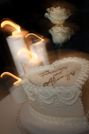 Heart-shaped white wedding cake inscribed Forever and Always surrounded by glowing candles and white roses.
