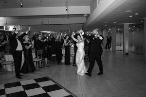 Black and white photo of bride and groom dancing at wedding reception with guests cheering on checkered dance floor.