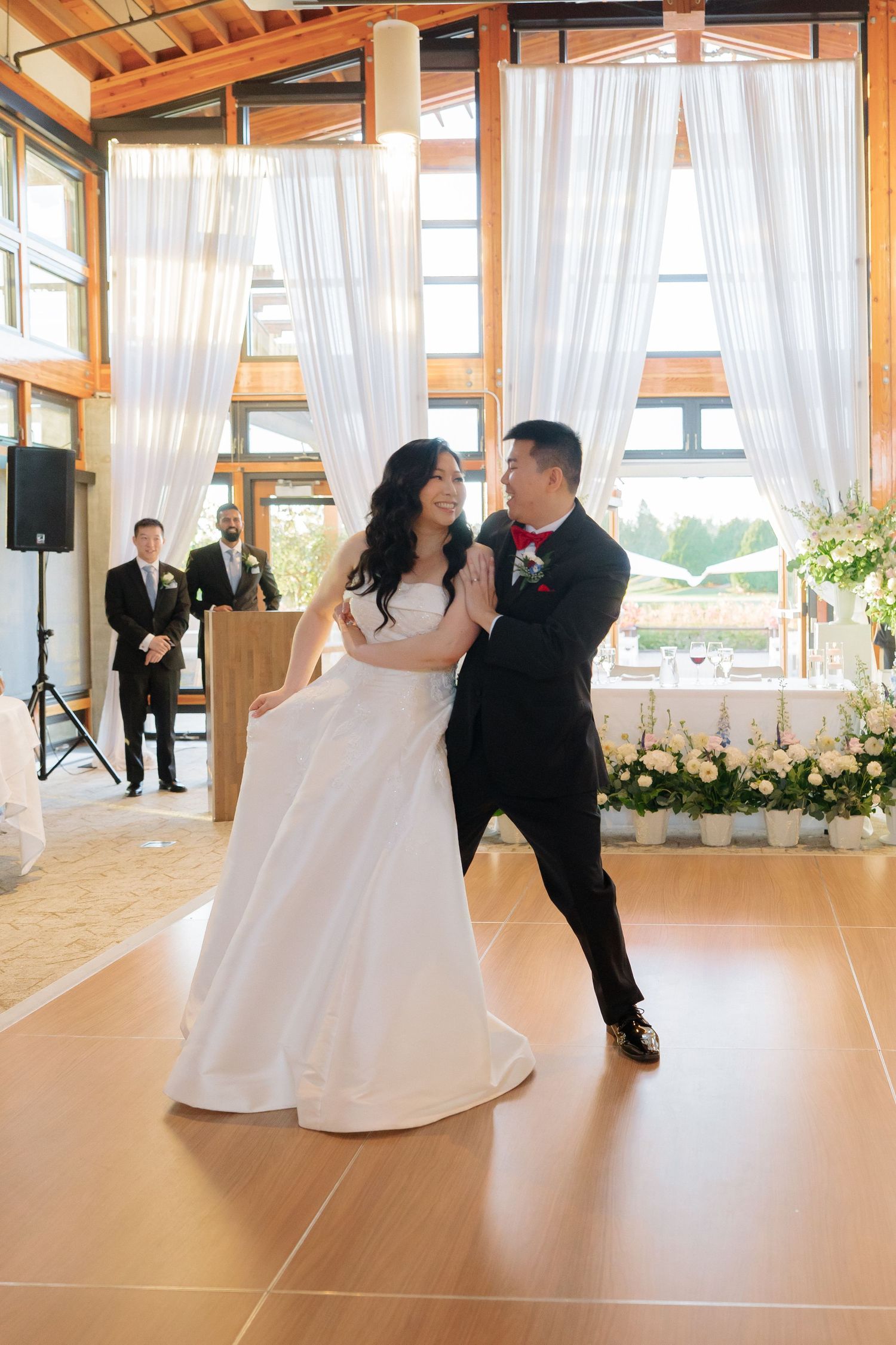 Bride in a white gown and groom in a black tuxedo share a first dance on a wooden floor in a sunlit venue with sheer white curtains.