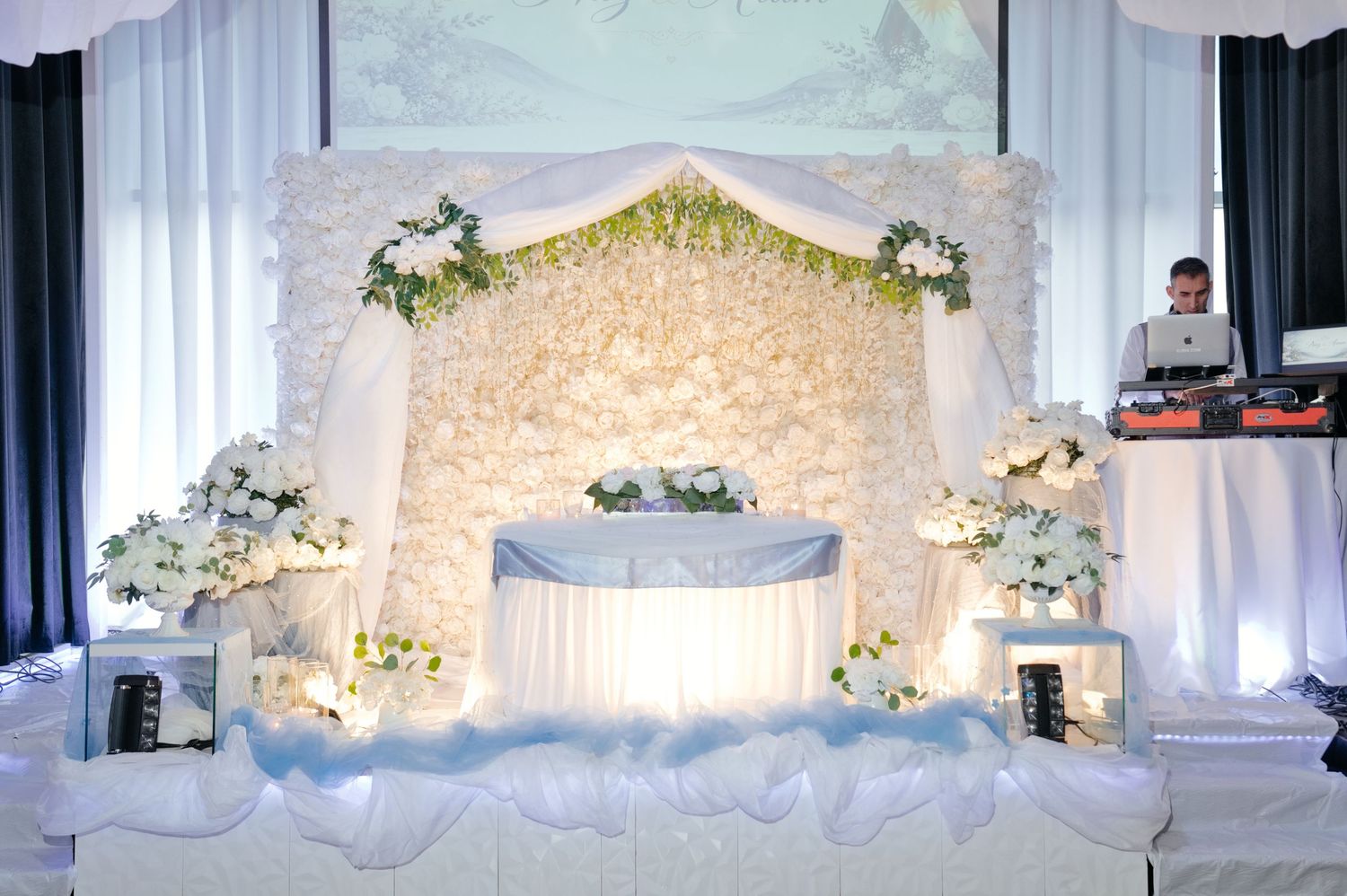 Elegant white wedding reception sweetheart table with floral backdrop, draped fabric and flower arrangements, with a DJ at his setup nearby.