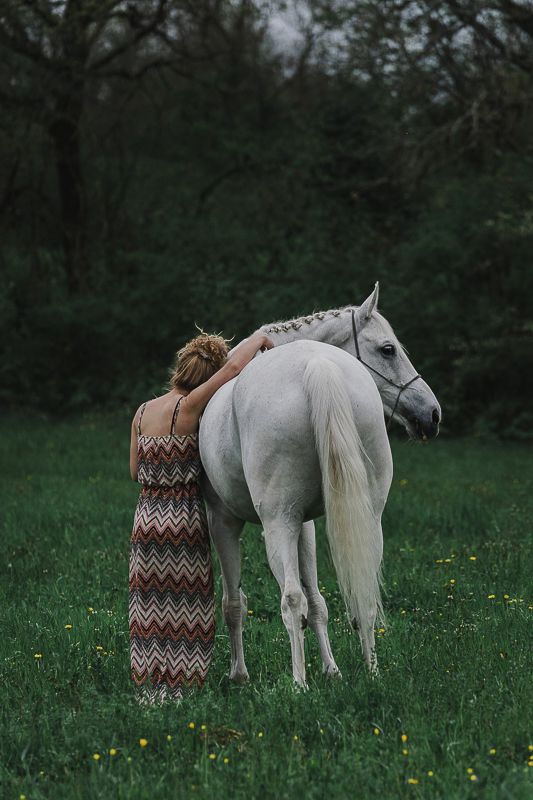 Woman in a chevron maxi dress leans against a white horse in a green grassy field near dark woods.