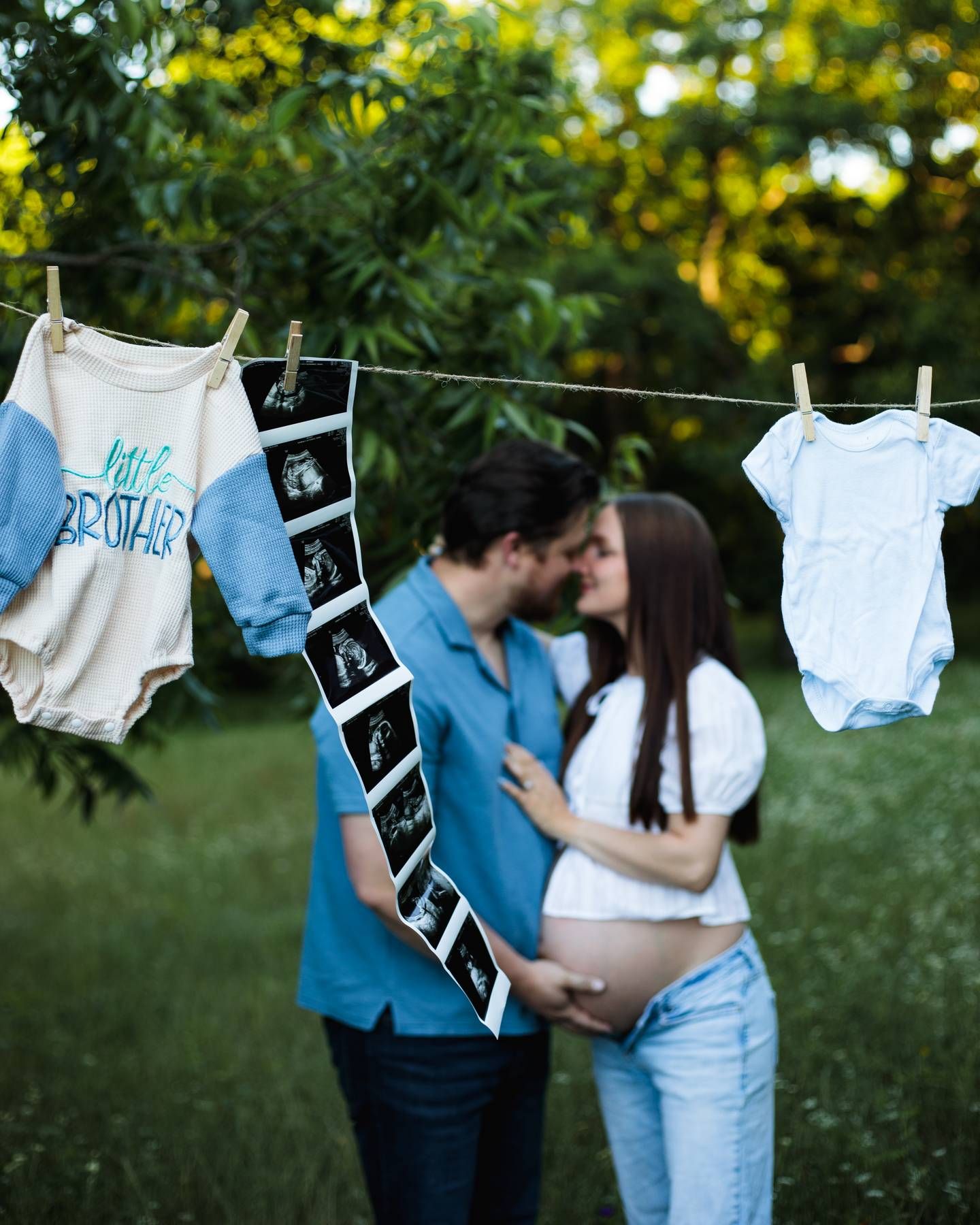 Expecting couple embrace beside a clothesline with baby onesies and ultrasound photos, one reading big brother, in a green park.