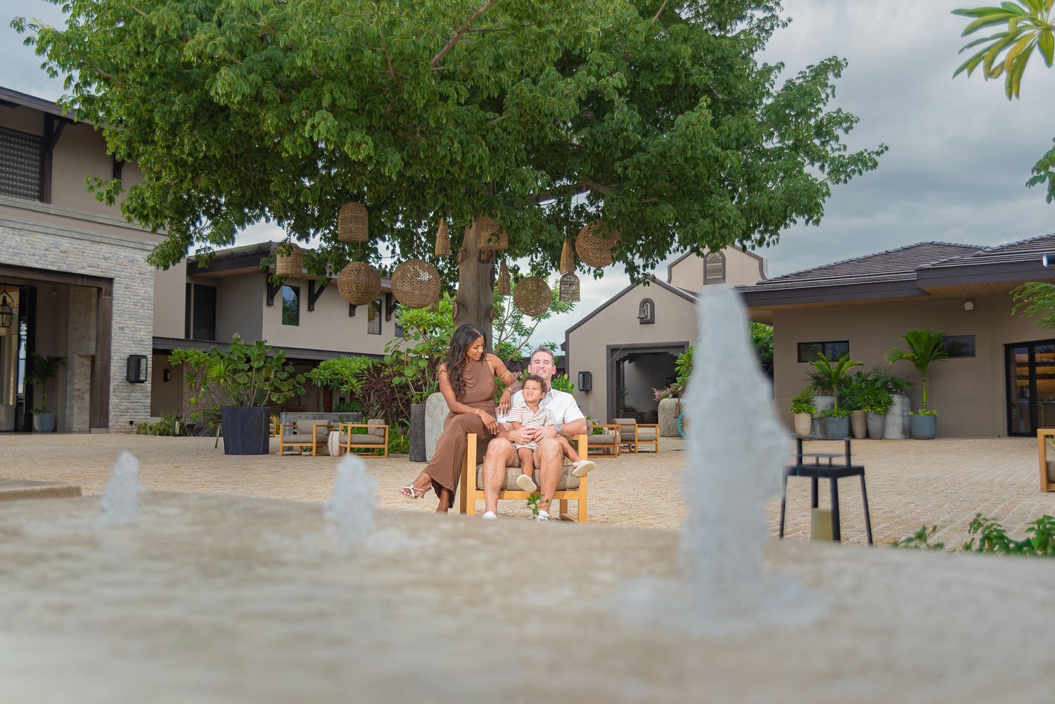 Two people sit on a bench under a large shady tree beside a water fountain in a sunny resort courtyard surrounded by modern buildings.