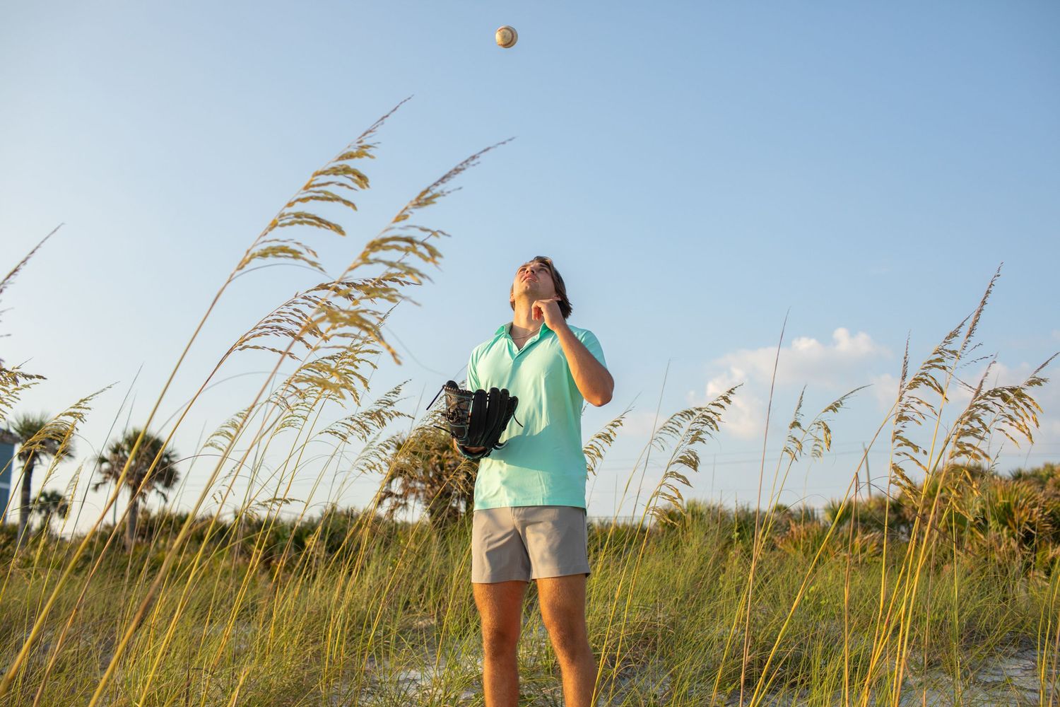 Woman in teal shirt tossing a ball on a sunny beach with sea oats and palm trees in the background.