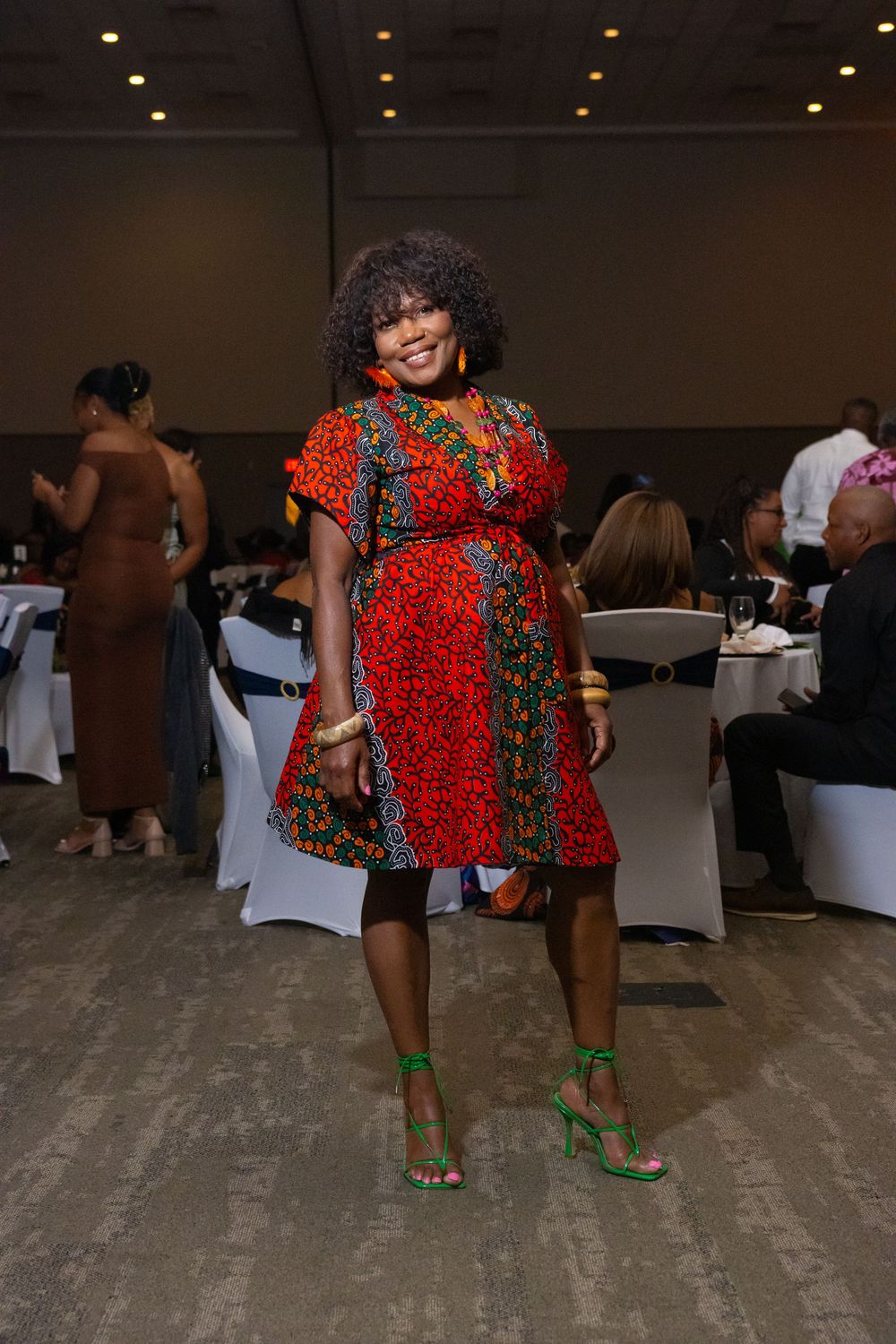 Woman in vibrant red African print dress and green heels smiles at an elegant indoor banquet event.