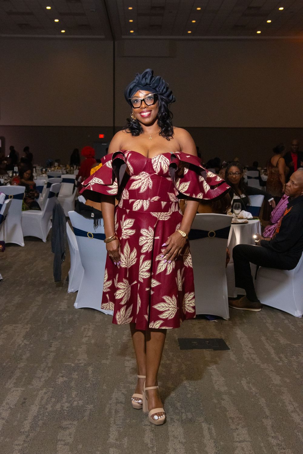 Woman in red floral off-shoulder dress and blue headwrap poses at elegant banquet hall event.