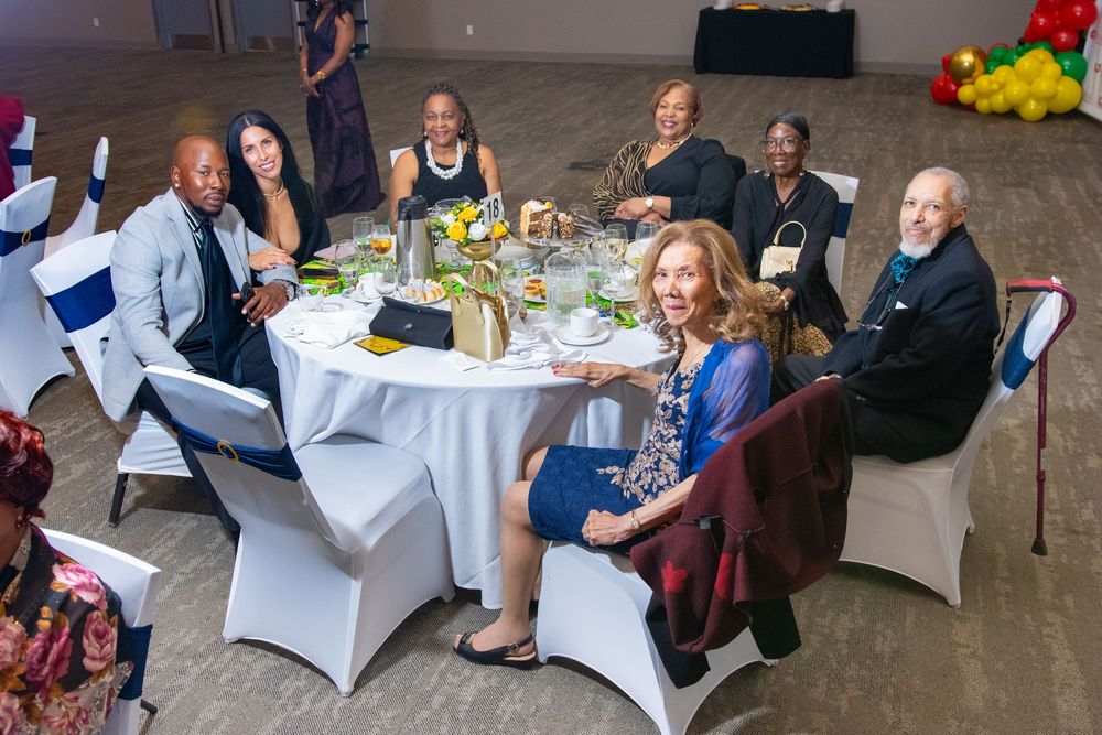 Diverse group of elegantly dressed guests smiling at a round banquet table decorated with yellow flowers at a formal event.