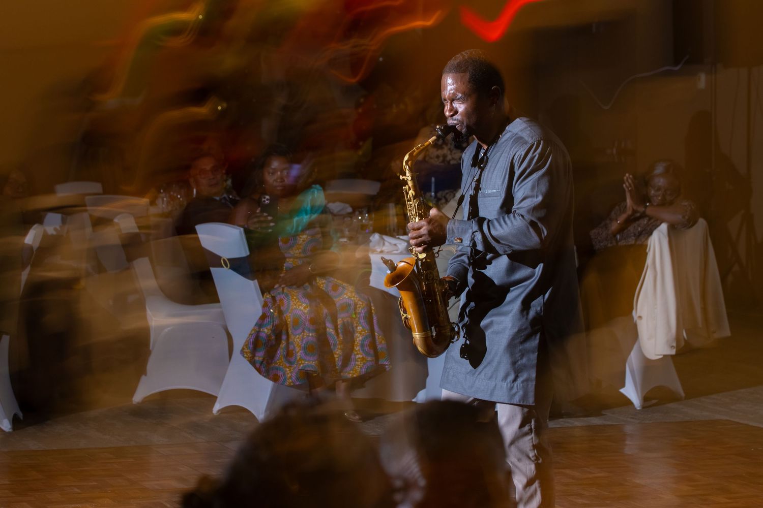 A saxophonist performs at an indoor event, with blurred guests and warm ambient lighting in the background.