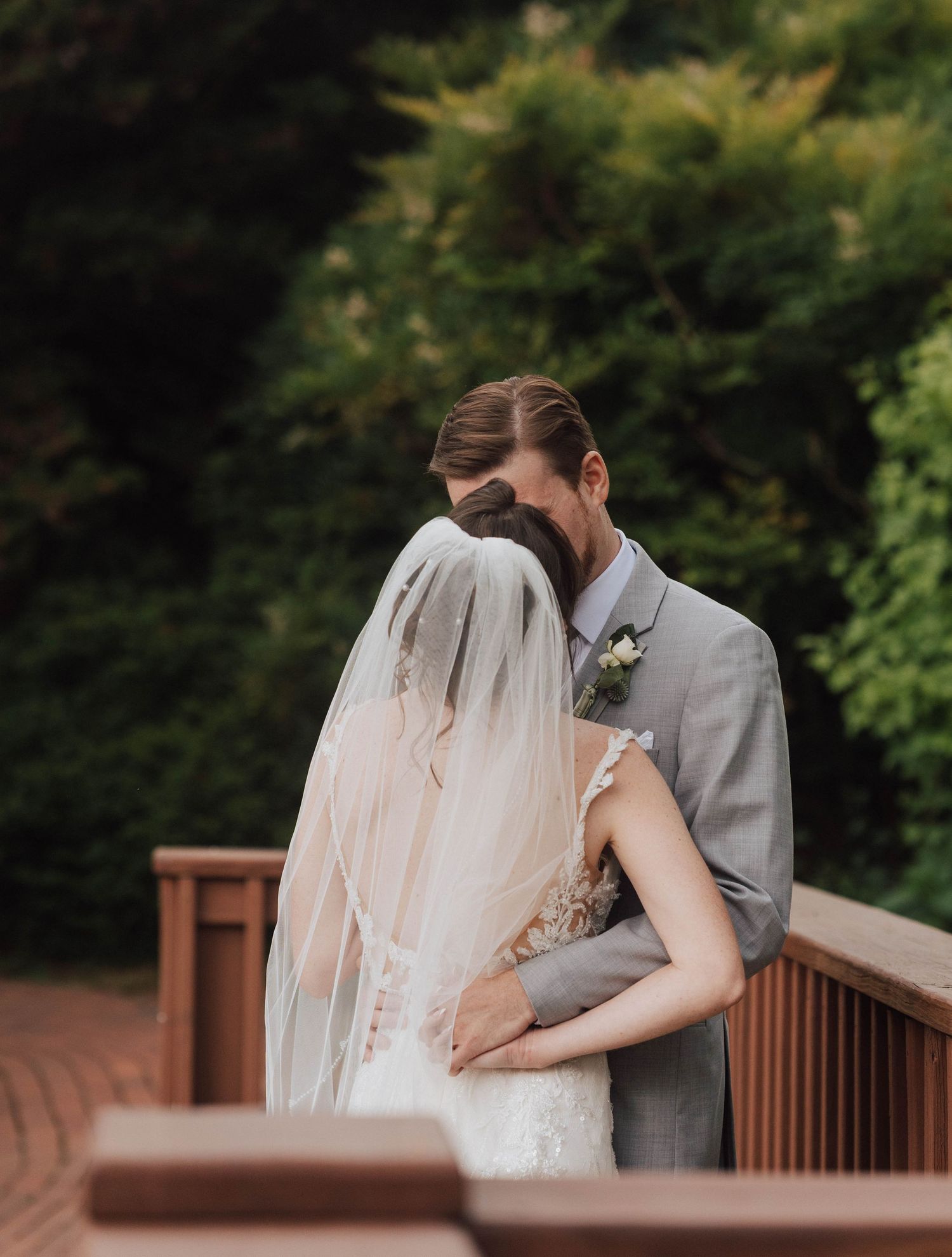 A bride and groom share an intimate embrace on a wooden deck surrounded by lush greenery on their wedding day.
