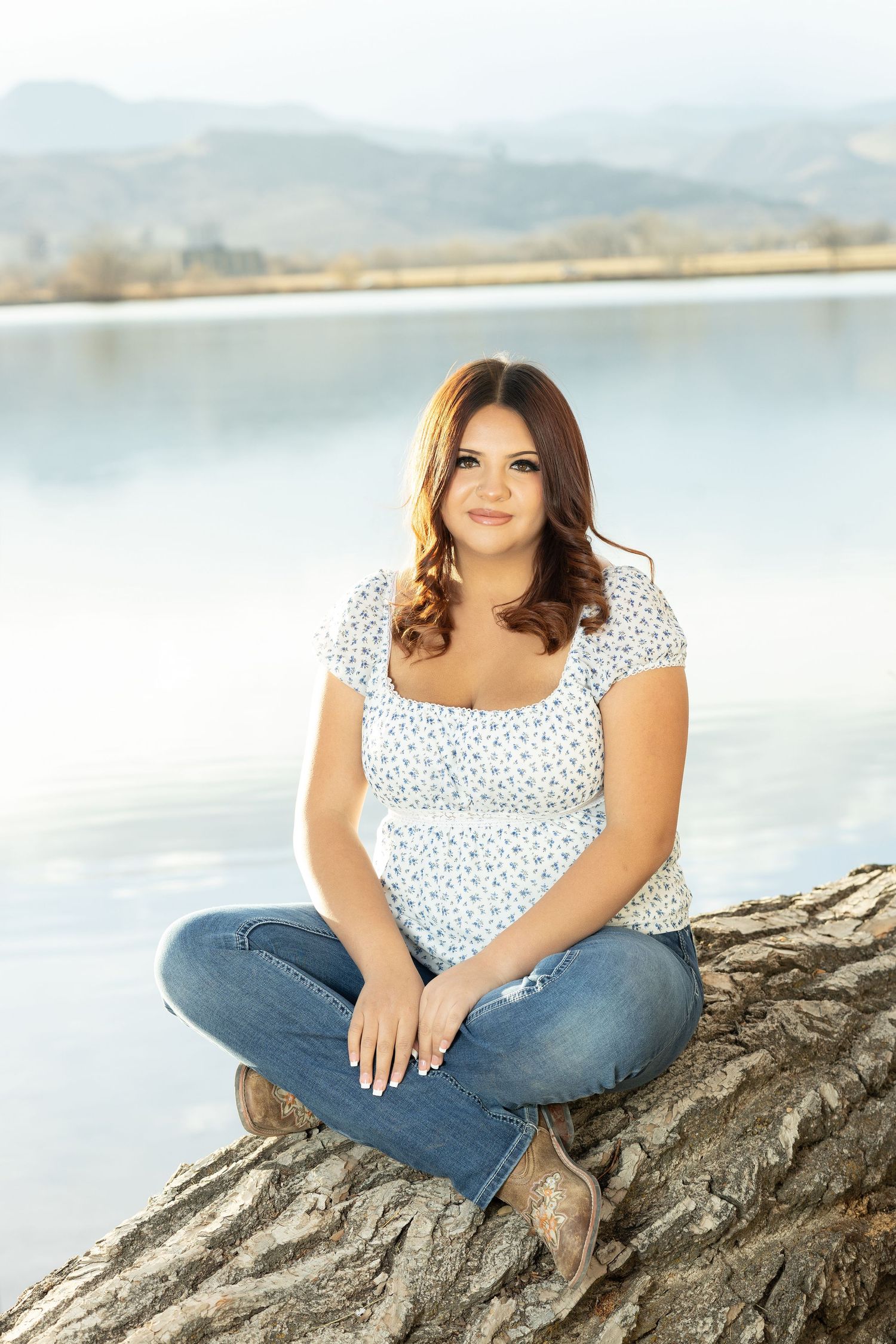 Young woman in white floral top and jeans sitting cross-legged on a rock by a serene lake with mountains in the background.