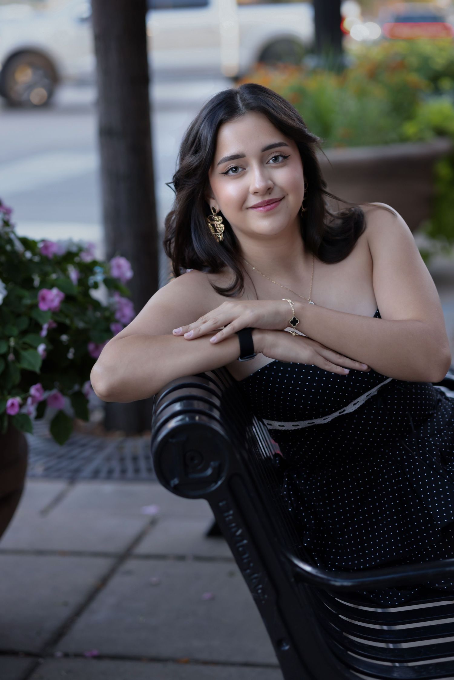 Young woman in black polka dot dress sitting on iron bench outdoors, wearing gold earrings and smiling.