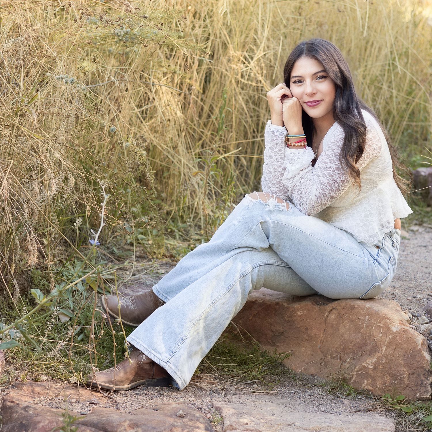 Young woman in floral top, light jeans, and brown boots sits on a rock surrounded by tall golden grass.