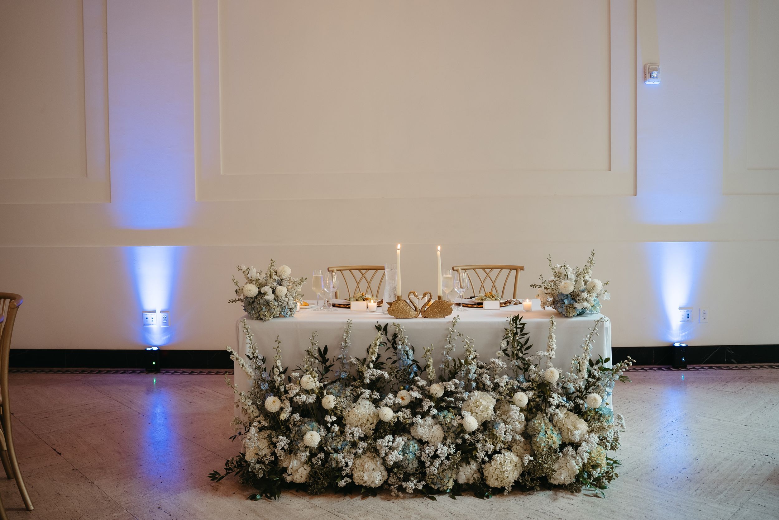 Elegant wedding sweetheart table adorned with white floral arrangements, candles, and blue uplighting in a ballroom.