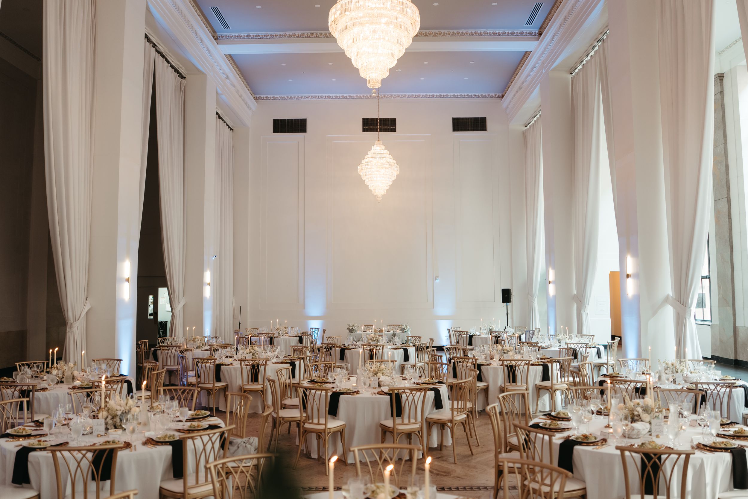 Elegant wedding reception hall with crystal chandeliers, white draping, round tables, and gold chiavari chairs in a grand ballroom.
