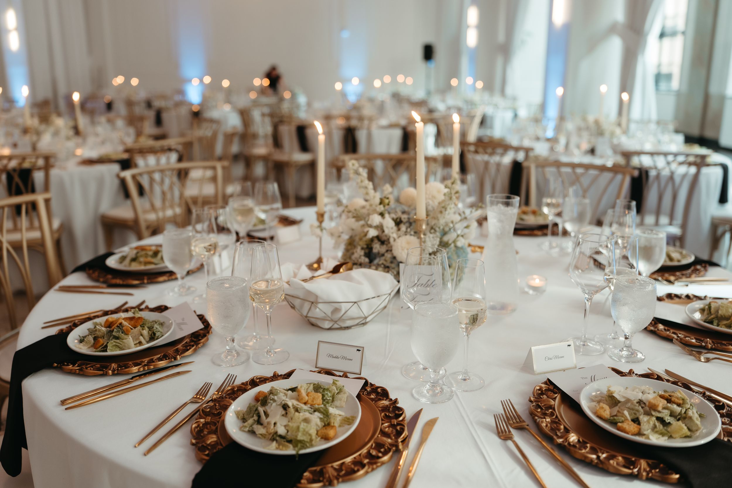 Elegant wedding reception table set with gold chargers, white linens, crystal glasses, candles, and floral centerpiece.