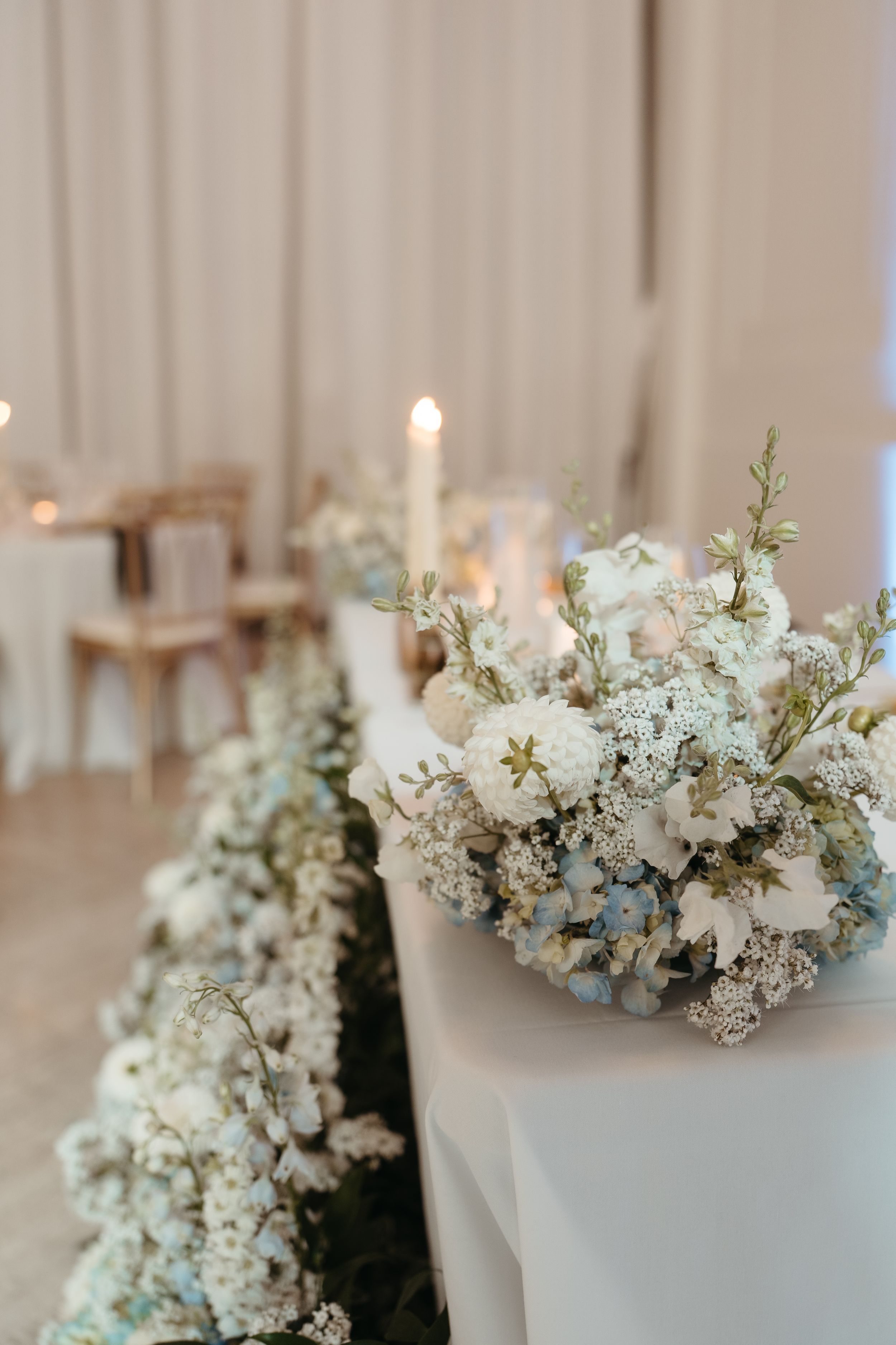 Elegant wedding table with white and blue floral arrangement, candles, and draped fabric backdrop.