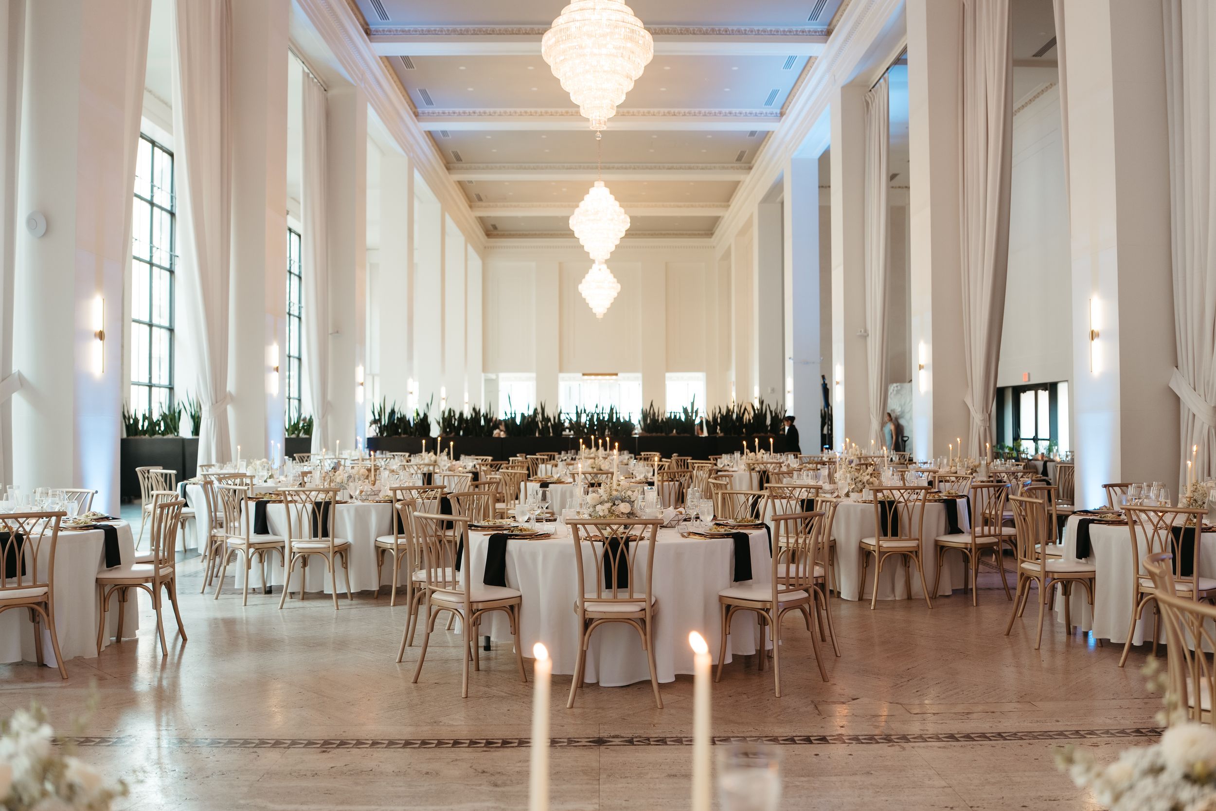 Elegant wedding reception hall with round tables, gold chairs, white linens, and geometric chandeliers in a grand ballroom.