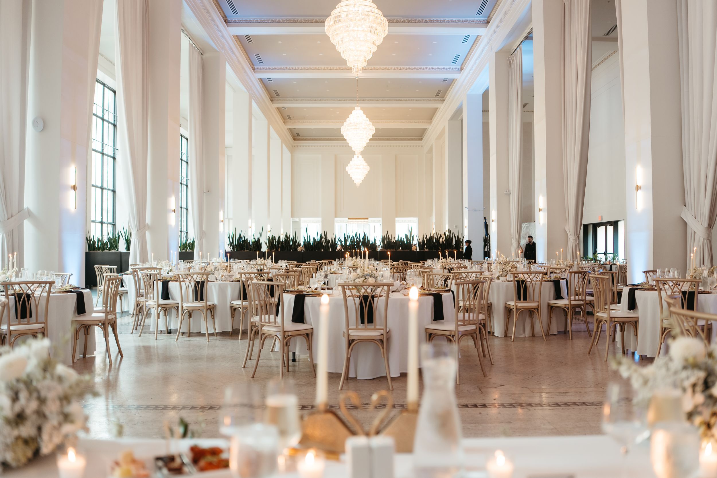 Elegant wedding reception hall with white columns, chandelier lighting, gold chiavari chairs, and neutral floral centerpieces.