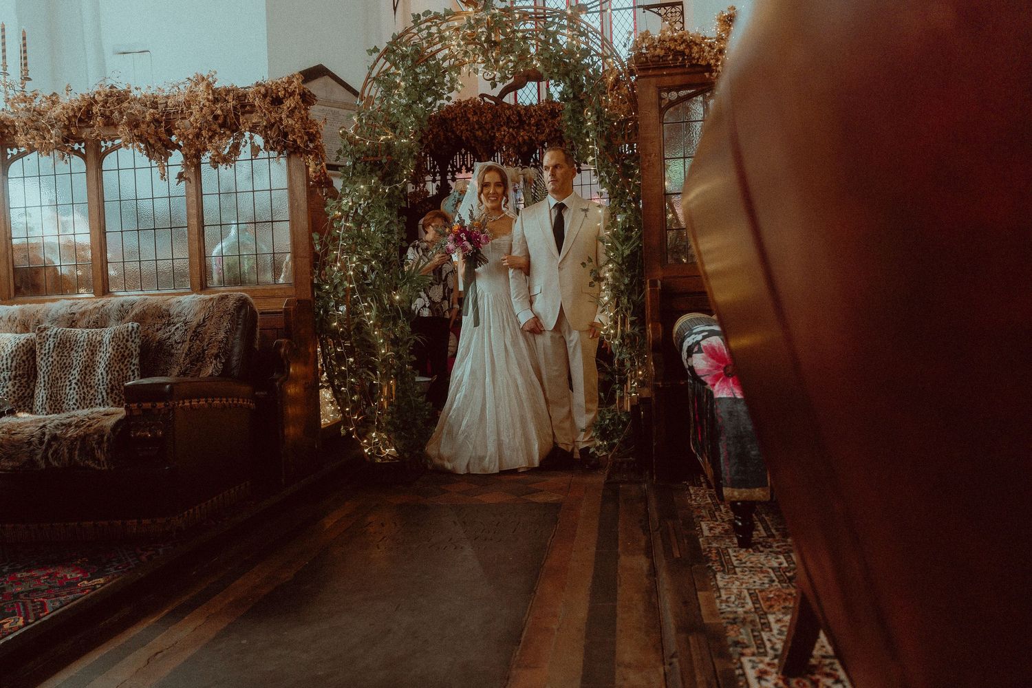 Bride and groom stand beneath a floral arch adorned with greenery and fairy lights during an elegant indoor wedding ceremony.
