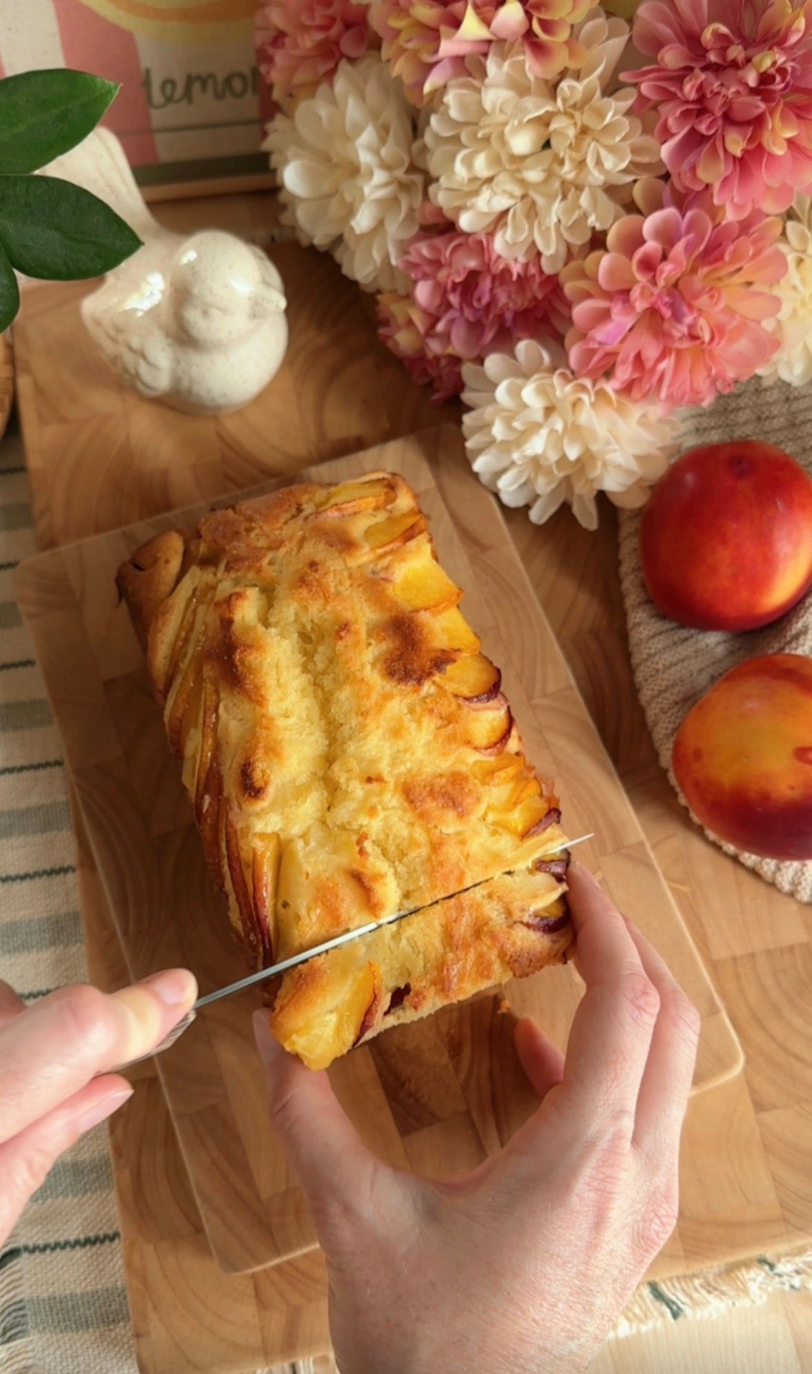 Sliced peaches arranged in overlapping rows in a parchment-lined loaf pan, ready for baking, with pink flowers in background.