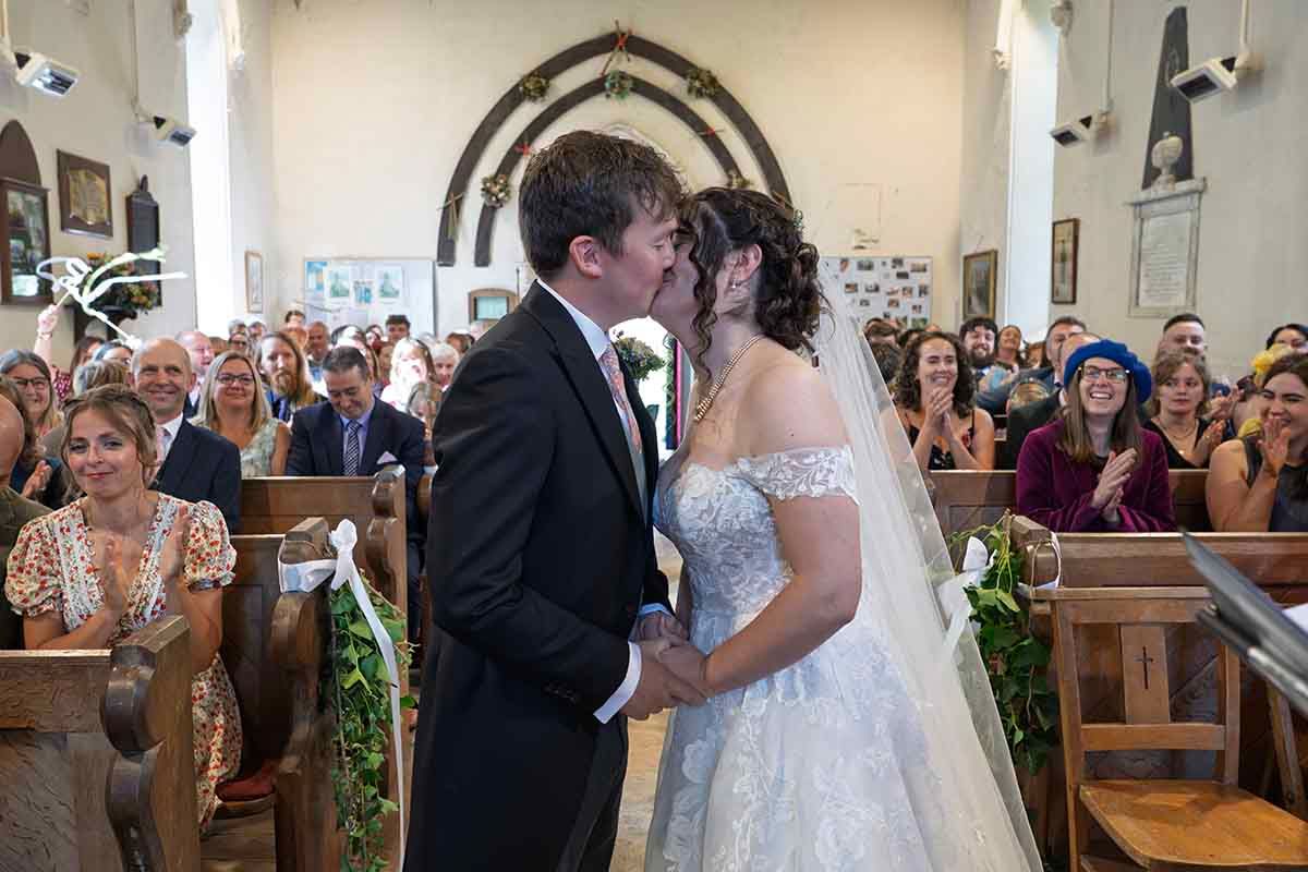 Bride and groom share first kiss at church wedding altar, surrounded by applauding guests in decorated pews.