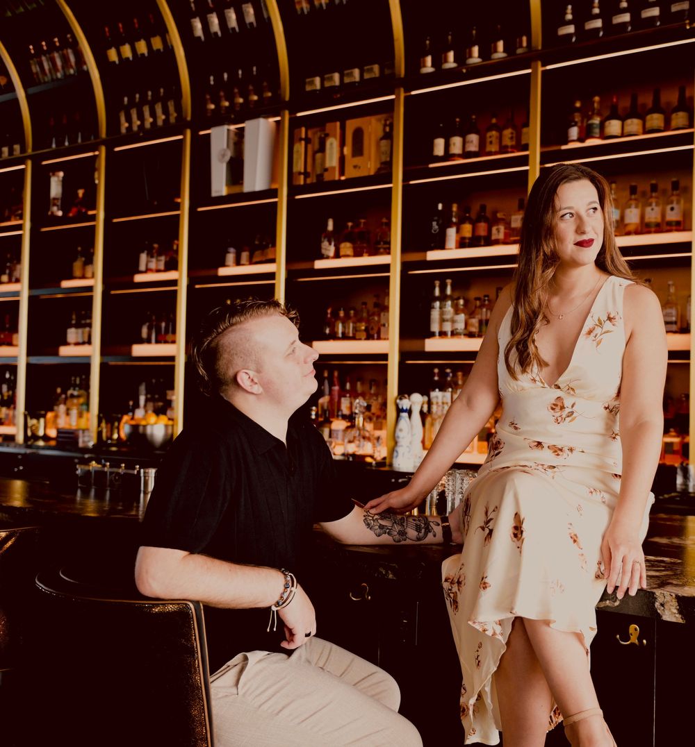 Couple at a bar, man seated looking up at woman in floral dress, shelves of bottles behind them.