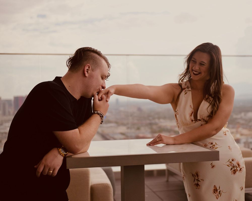 A couple shares a romantic moment on a rooftop balcony, man kissing womans hand while she laughs joyfully.