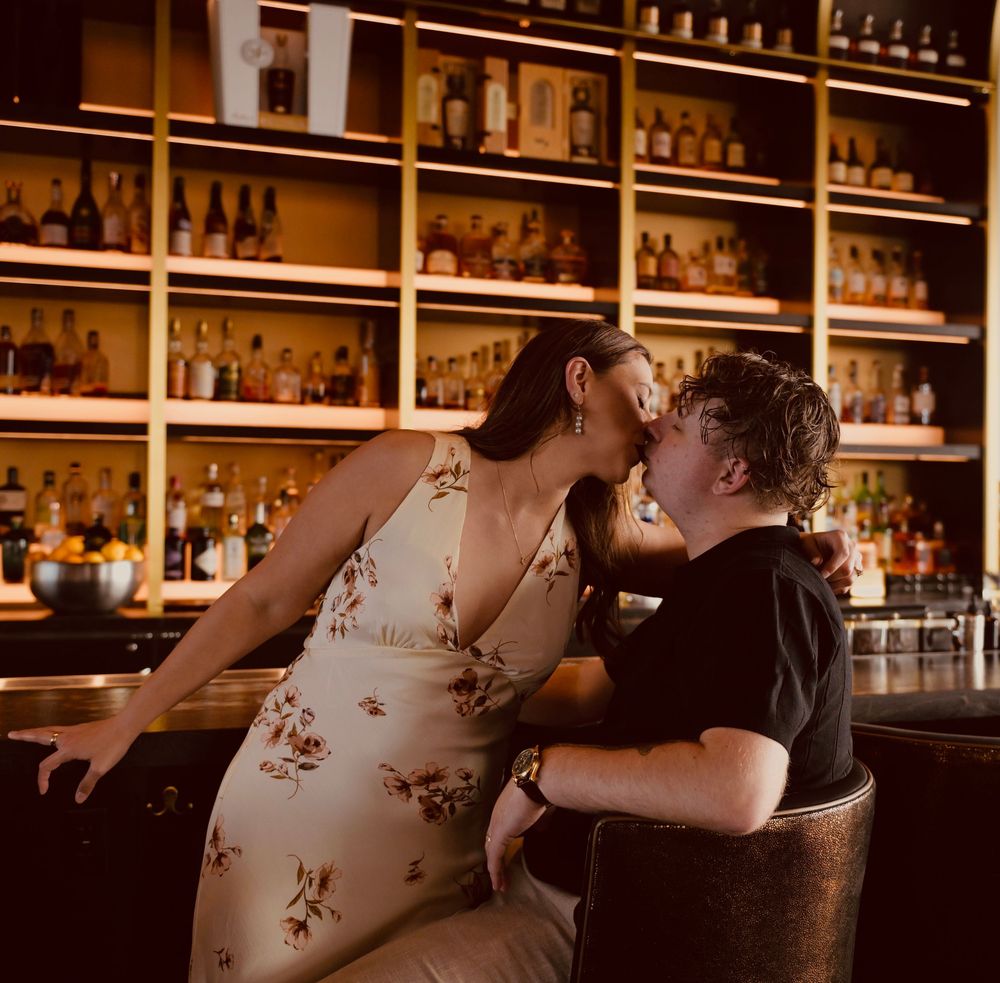 Couple sharing a romantic kiss at a warmly lit bar with shelves of liquor bottles in the background.
