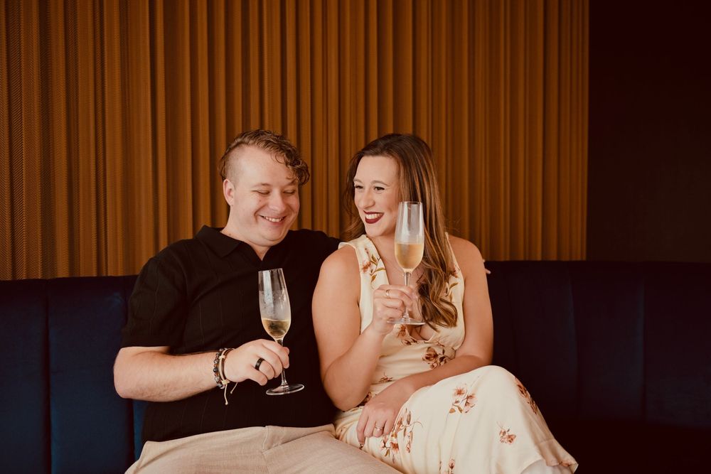 Couple laughing and toasting with champagne glasses on a blue velvet sofa in a warmly lit venue.