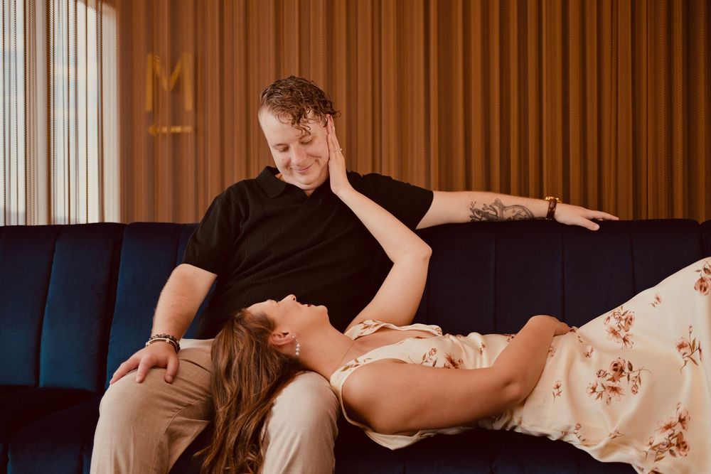Couple relaxing on blue velvet sofa, woman lying across man's lap laughing in stylish hotel lounge.