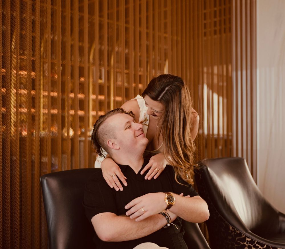 Couple embracing tenderly on a leather chair against warm wooden slat wall backdrop.