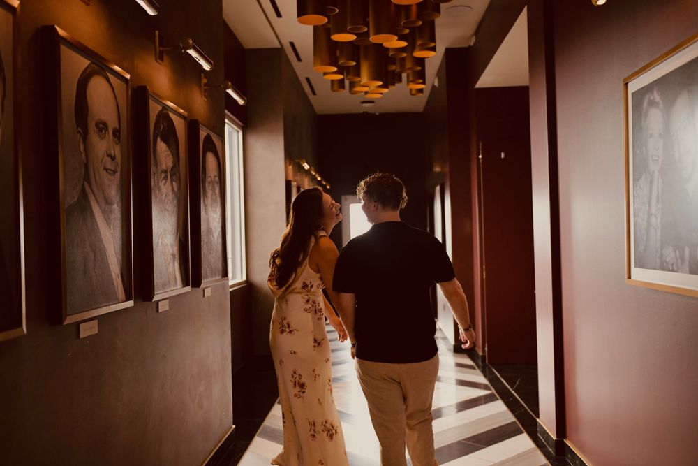 Couple walking hand in hand down an elegant hotel hallway with portrait artwork and warm ambient lighting.