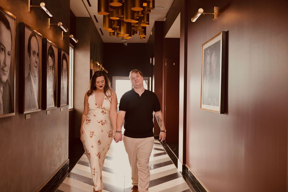 Couple holding hands walking down an elegant restaurant hallway with framed portraits and warm lighting.