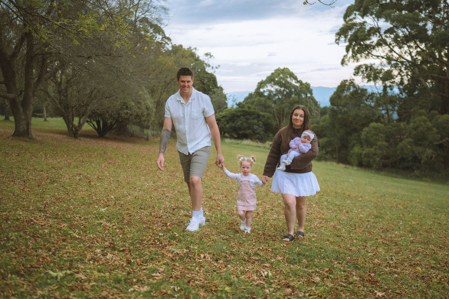 Family of four walking on grass in a park, parents holding toddler's hands while mother carries a baby.