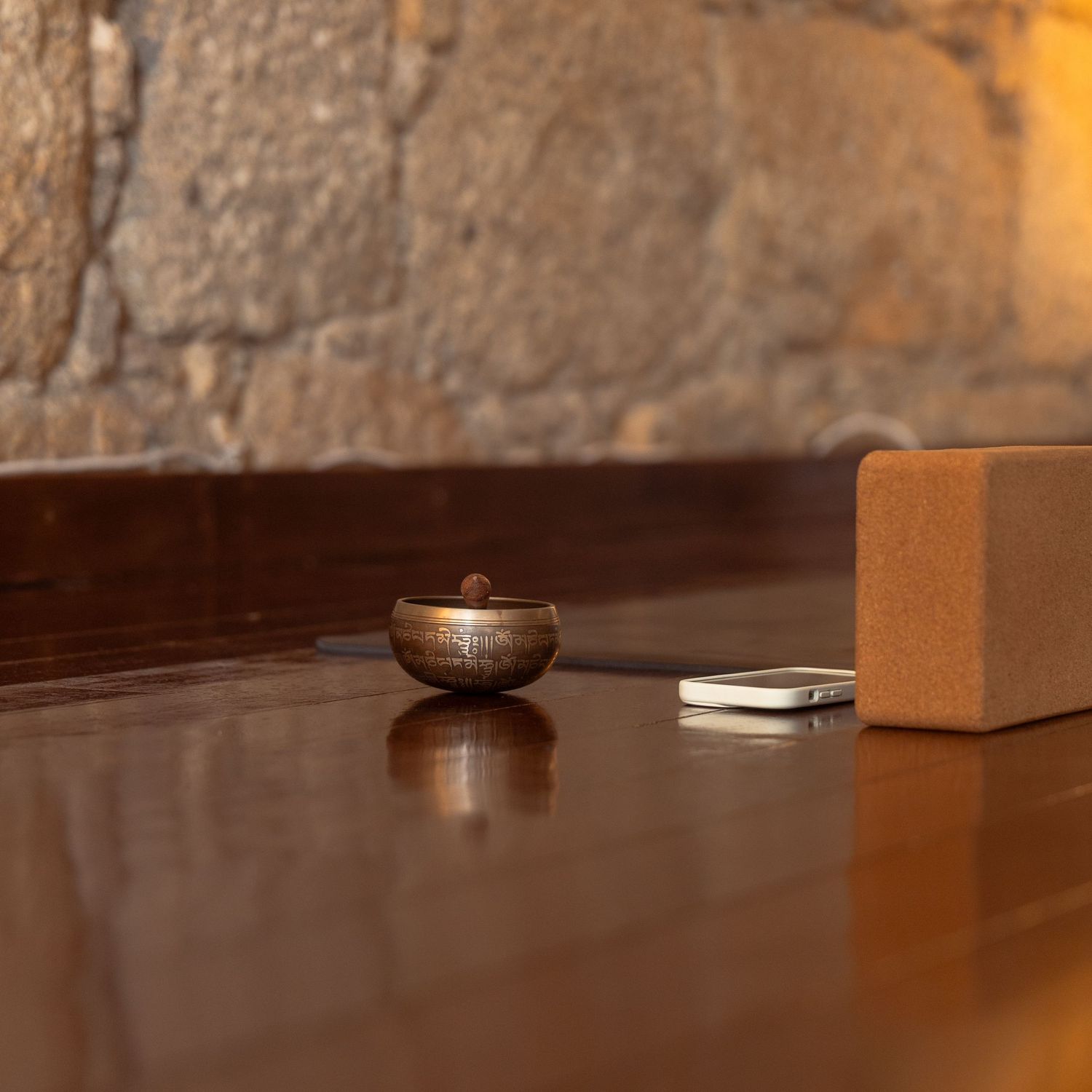 A Tibetan singing bowl and cork yoga block on a wooden floor against a rustic stone wall.