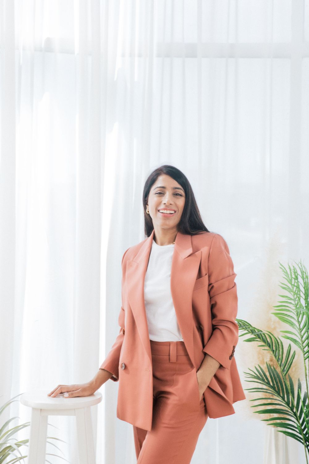 Professional woman in coral business suit poses confidently against white backdrop with palm plant.