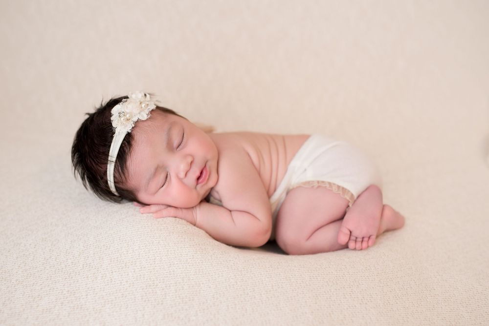 Smiling newborn baby girl sleeping peacefully on a cream blanket, wearing a white floral headband and bloomers.