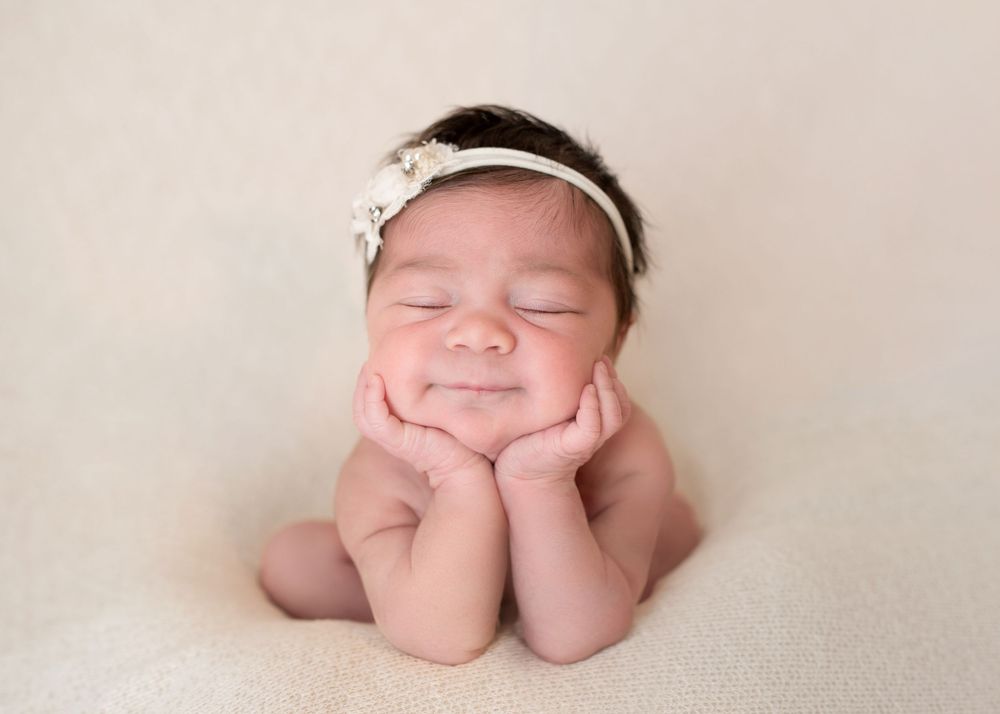 Smiling newborn baby girl with floral headband resting chin on hands in classic froggy pose on cream background.