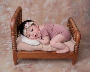 Newborn baby in pink romper and floral headband posed on a miniature wooden bed with a white pillow.